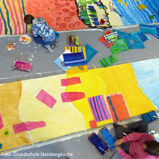 Zwei Kinder malen großflächig ihre auf dem Boden liegenden Kunstwerke